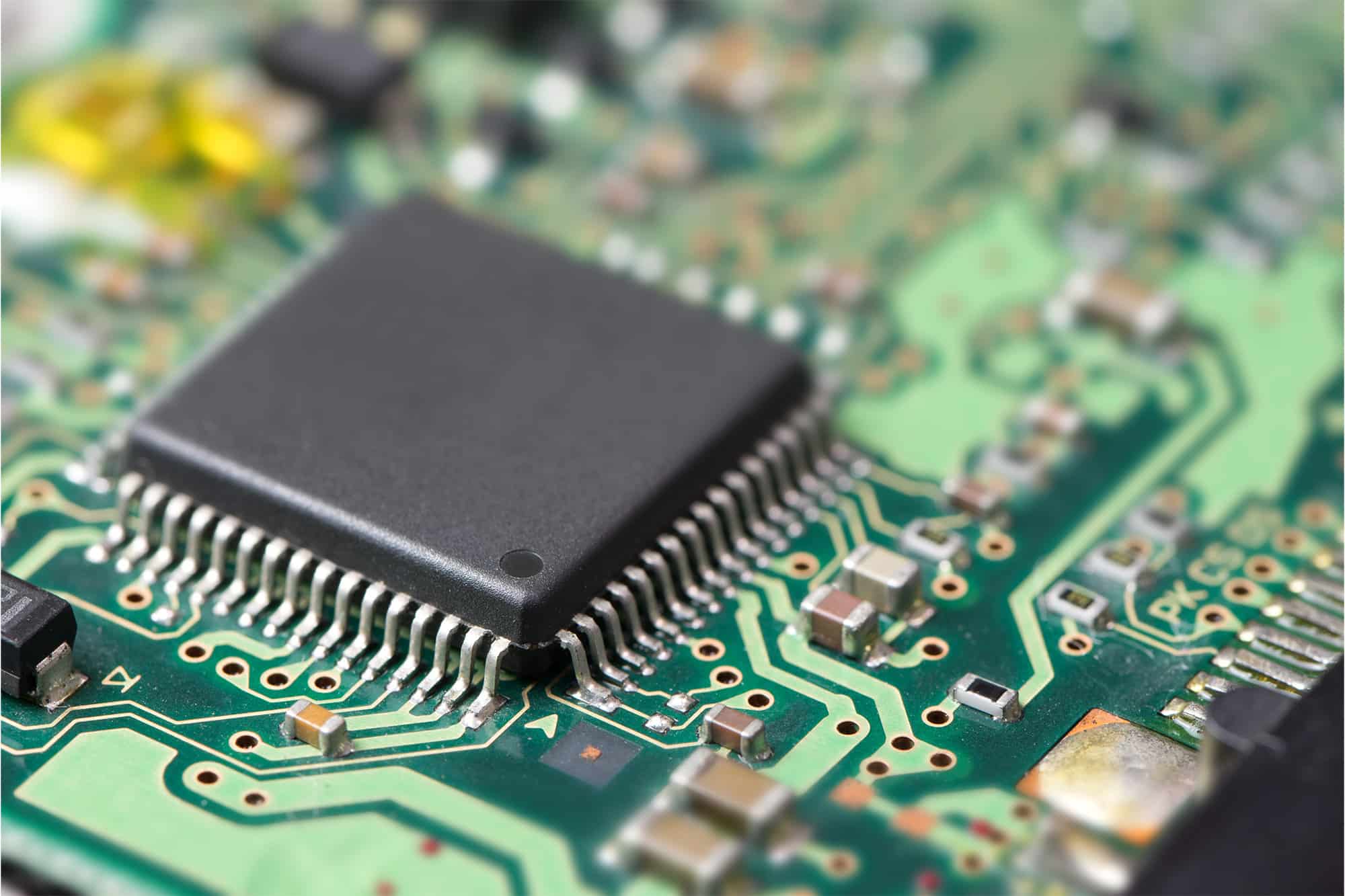 Close-up of a green circuit board with a large black integrated circuit and numerous silver pins soldered to the traces.